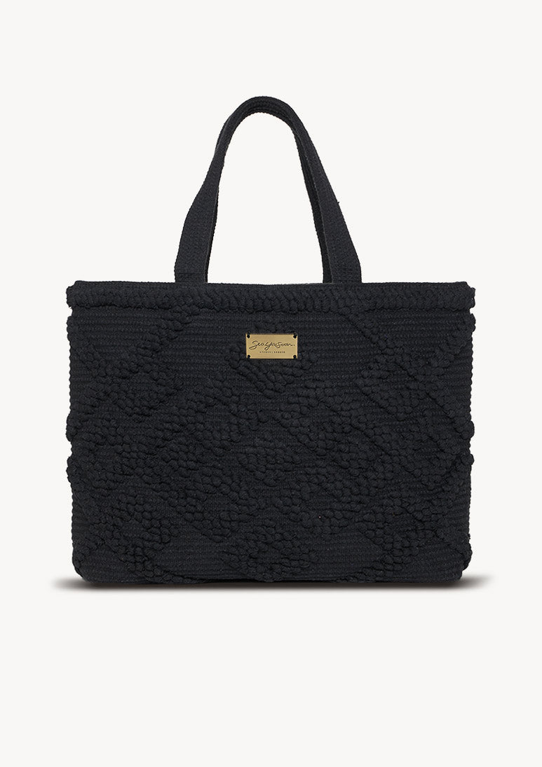 A black cotton tote bag with a unique pom pom design, thick handles, and a canvas-lined interior with a zipper pocket.